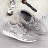 Scarpe da ginnastica grigie e argento su sfondo in legno chiaro, con occhiali da sole, orologio e tappeto bianco. Design moderno e casual.