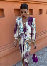 Donna con abito lungo a fantasia astratta viola e beige, borsa viola, orecchini dorati, sfondo arancione. Moda elegante, street style.